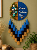 A decorative hanging wall piece with a split design: one half is a blue semicircle with white personalized text reading 'Karan Nishma B1701' and a small heart, the other half is a wood-grain circle with greenery and blue/white flowers. Below hang tassels in blue, black, and tan on a green wall, with dried flowers, a candle, and a plant nearby.