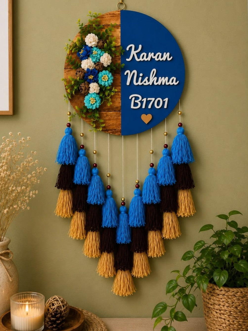 A decorative hanging wall piece with a split design: one half is a blue semicircle with white personalized text reading 'Karan Nishma B1701' and a small heart, the other half is a wood-grain circle with greenery and blue/white flowers. Below hang tassels in blue, black, and tan on a green wall, with dried flowers, a candle, and a plant nearby.